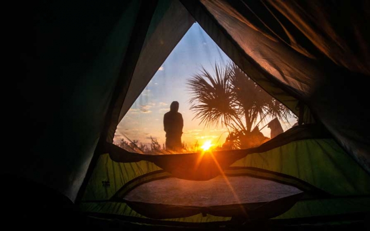 From inside of a tent, the sun is rising on the horizon. A person stands outside the tent, looking towards the sunrise.